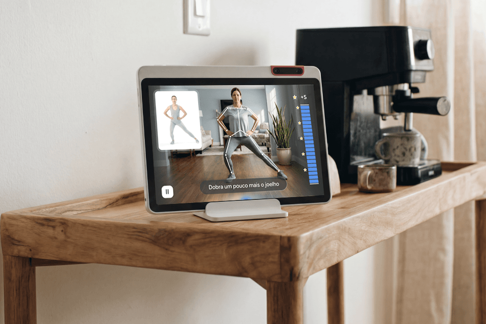 A tablet on a wooden table displays a workout video with a woman exercising. The screen shows exercise stats and instructions in Portuguese. A coffee machine and a jar are also on the table.
