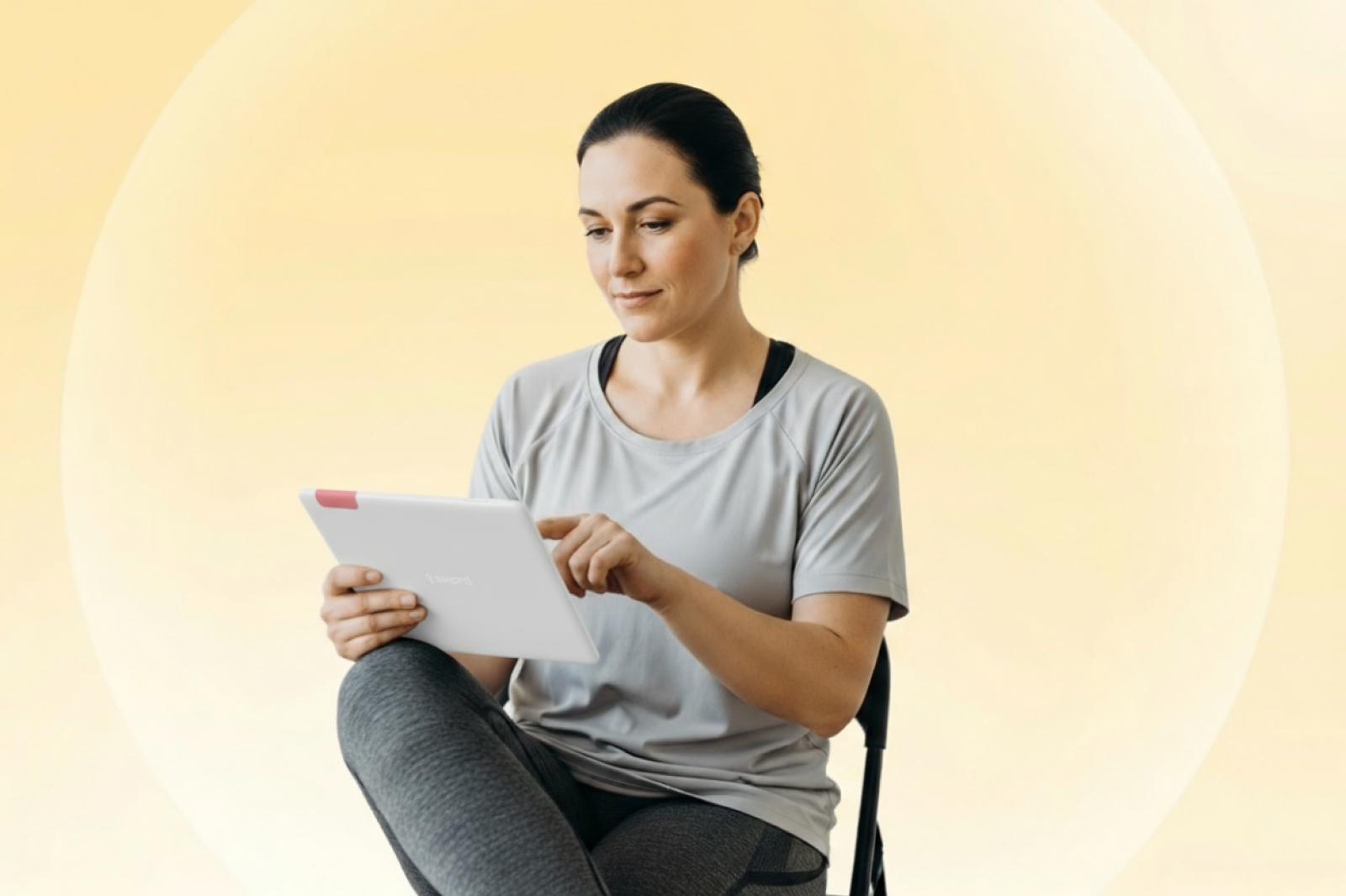 Woman in casual sportswear sitting and using a tablet against a soft yellow background.