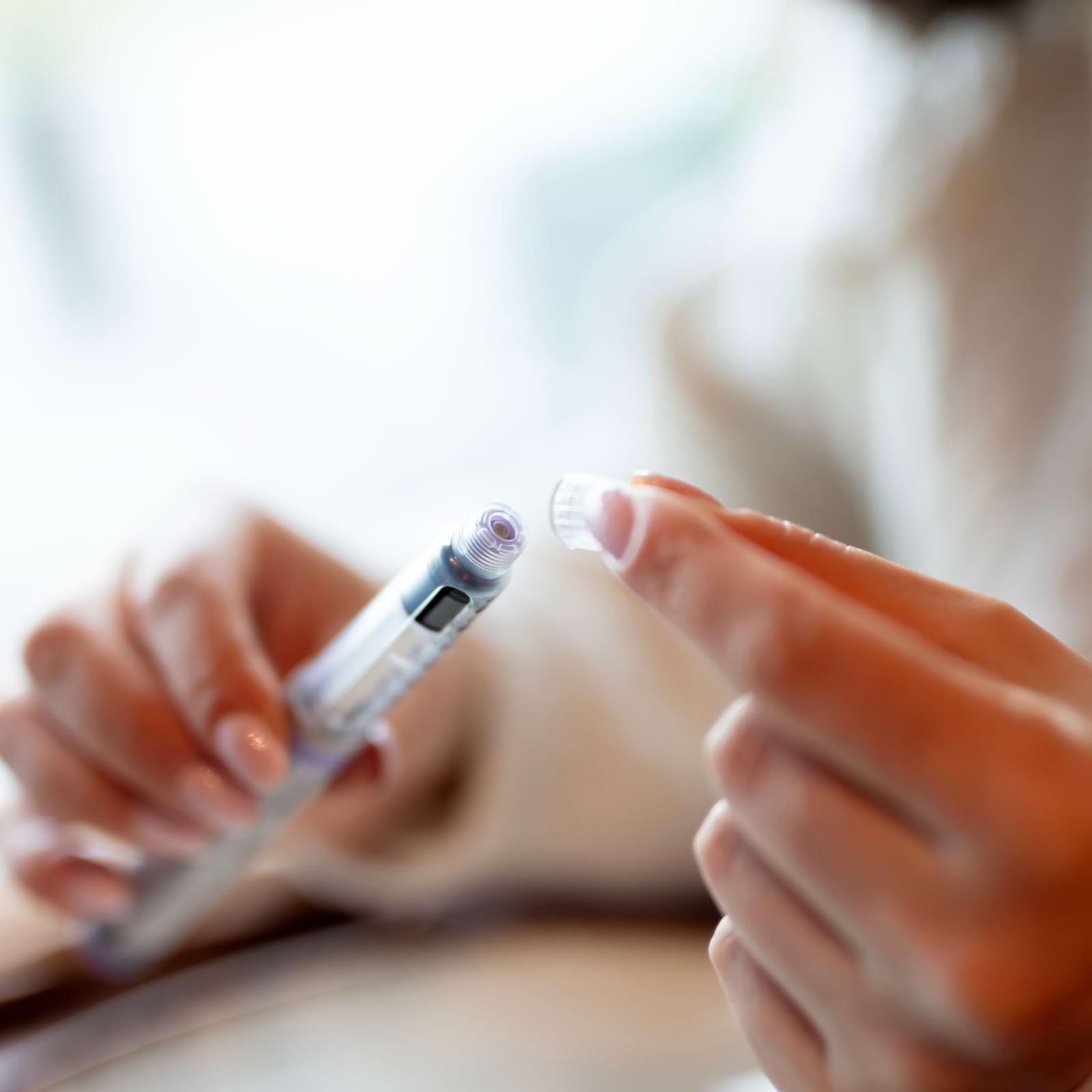 Close-up of hands preparing an insulin pen, with one hand removing the cap. The background is softly blurred.