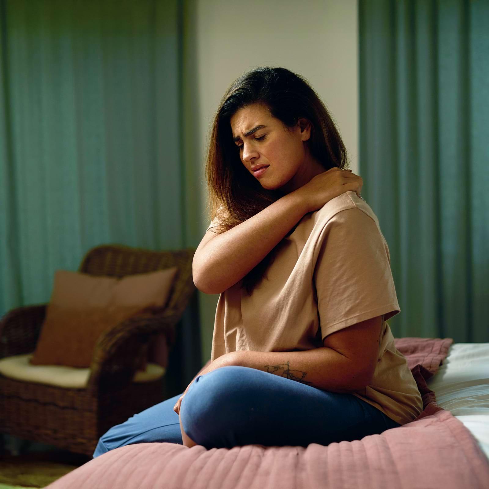 Woman sitting on a bed, holding her neck with a pained expression. She wears a beige shirt and blue pants. Room features wicker chair and curtains.