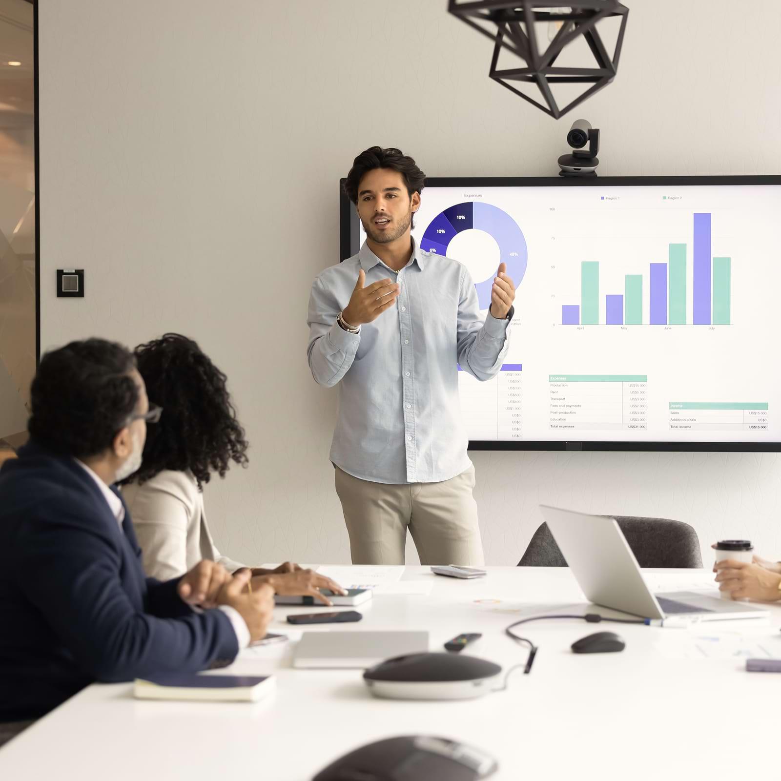 Man presenting data on a screen to colleagues in a conference room. Charts and graphs are visible in the background.