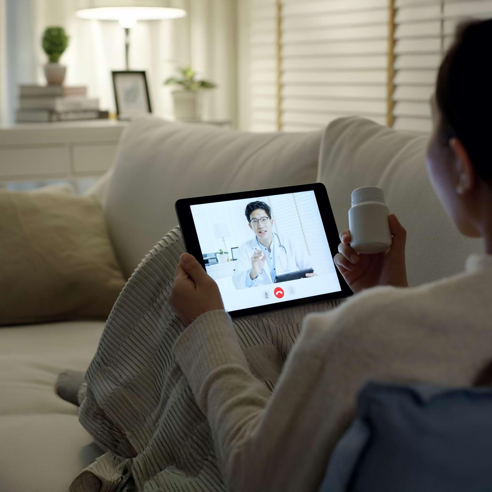 Person on a couch video chatting with a doctor on a tablet, holding a pill bottle; a lamp and decor in the background.