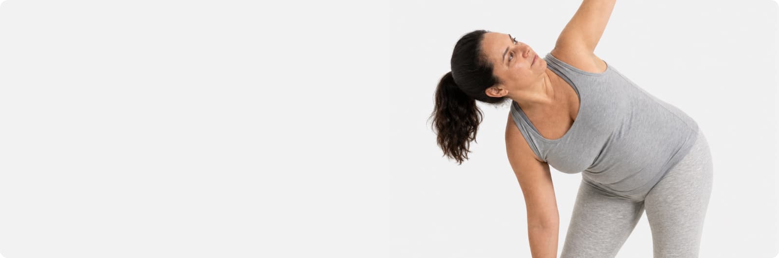 A woman in grey athletic wear stretches with one arm reaching upward and the other toward her foot, standing against a plain light background.