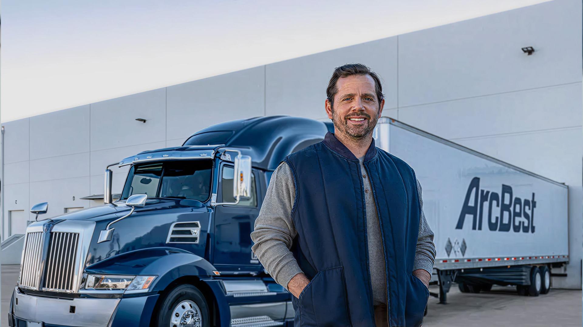 Smiling man standing in front of a blue semi truck and white ArcBest trailer outside a warehouse.