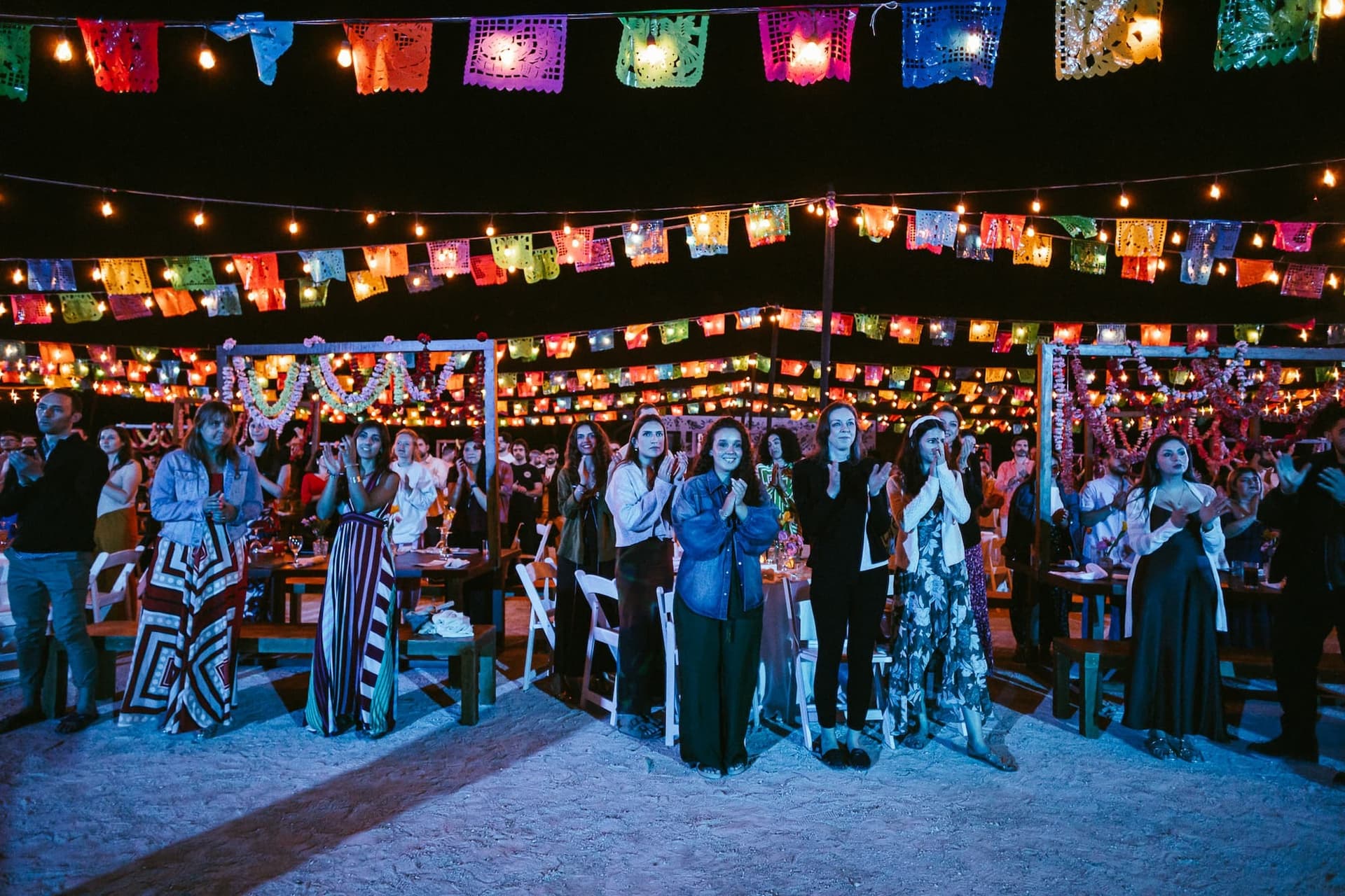 A group of people stand under vibrant, colorful string lights and flags at an outdoor event, clapping and celebrating on a sandy surface.