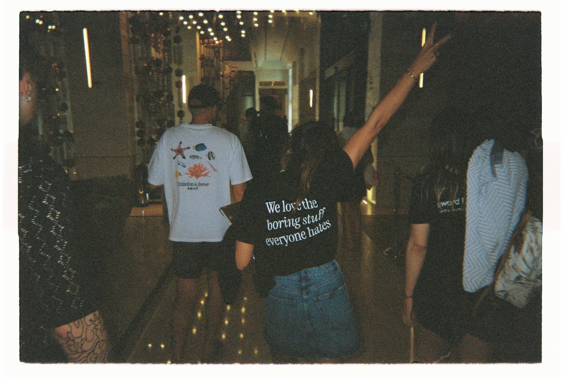 Group of people walking in dim lighting, one person facing away with a shirt that reads "We love the boring stuff everyone hates," raising a peace sign.