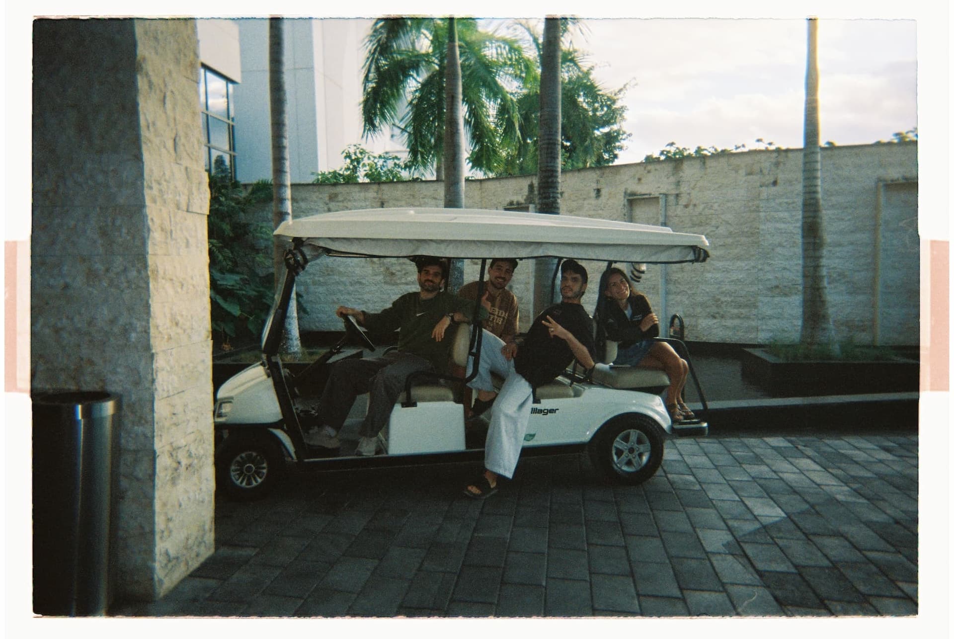 Four people seated on a golf cart parked on a paved path with palm trees and a building in the background.