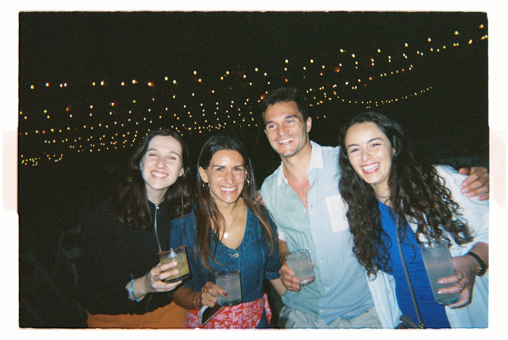 Four people smiling at an outdoor nighttime gathering with string lights above them.