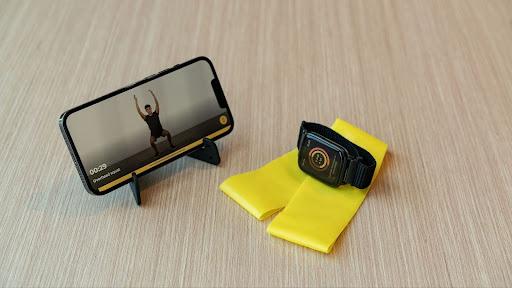Smartphone on a stand displaying a workout video, next to a smartwatch and a yellow resistance band on a wooden surface.