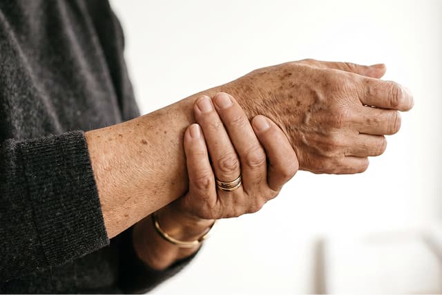 A person wearing a dark sweater holds their wrist with their other hand, showing signs of age such as wrinkles and age spots. They are wearing a gold ring and bracelet. The background is softly blurred.