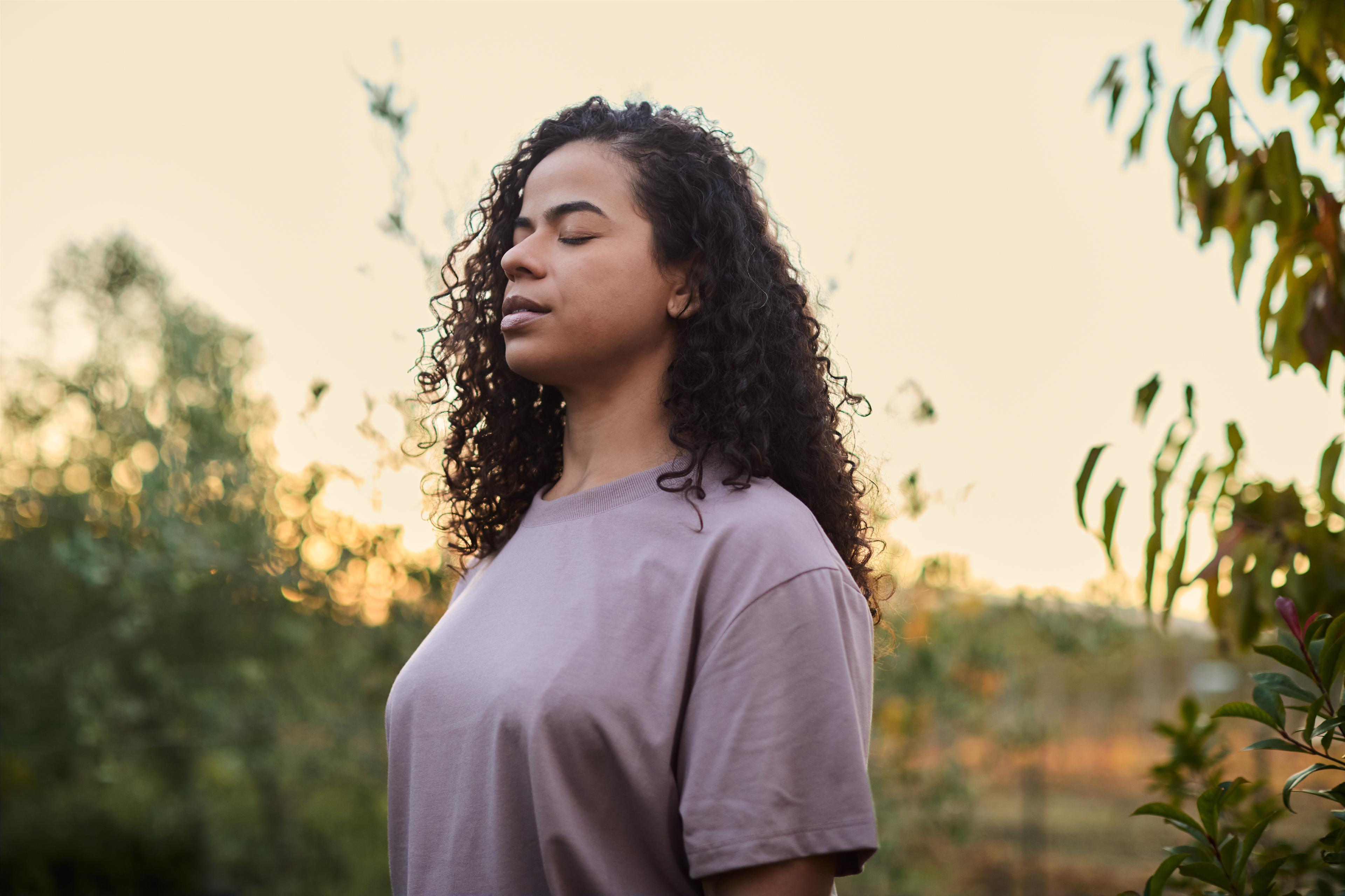 Woman with curly hair stands outdoors with eyes closed, wearing a purple shirt, surrounded by greenery and a soft sunset sky.
