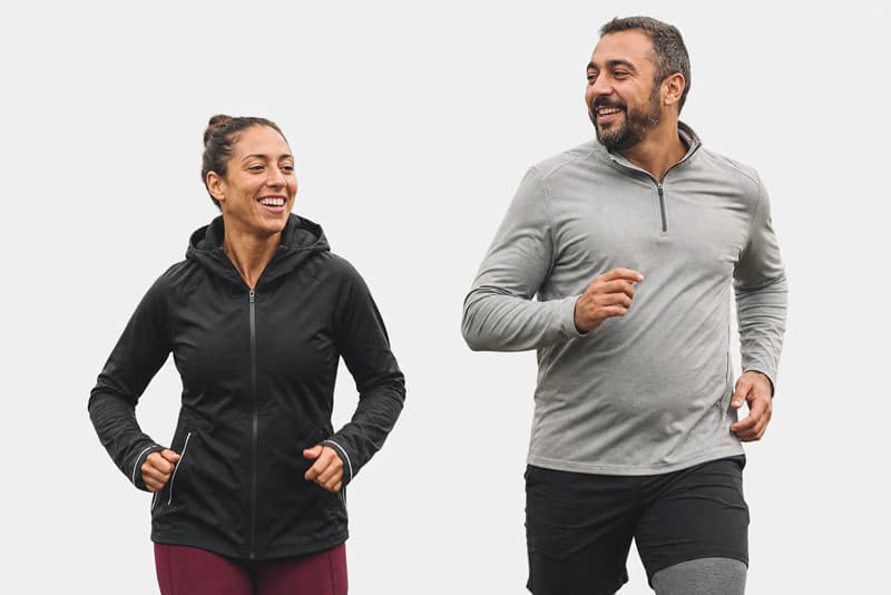 A woman and a man wearing activewear are jogging side by side, smiling at each other against a plain white background.