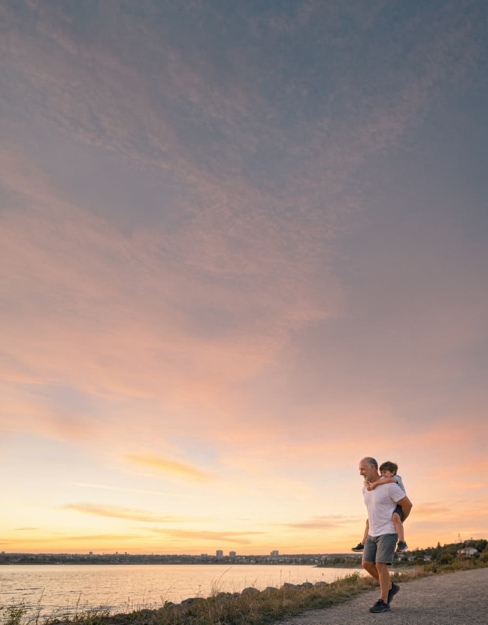 A man gives a child a piggyback ride along a waterfront path at sunset, with a wide sky filled with soft orange and pink clouds.
