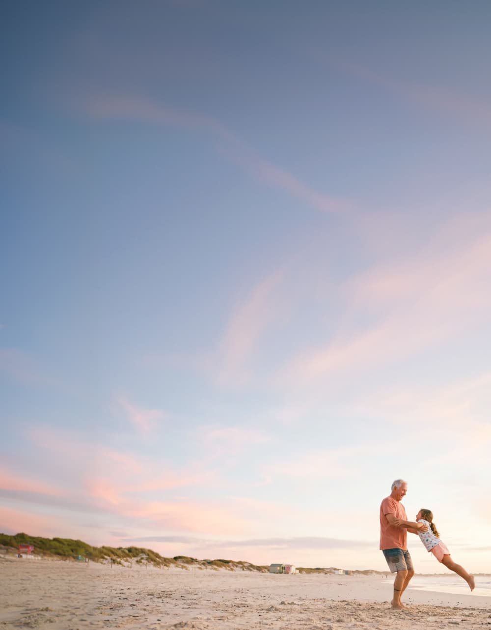 A man spins a child by the arms on a sandy beach at sunset, with a wide sky above them painted in soft pink and blue hues. Dunes and greenery are visible in the background.