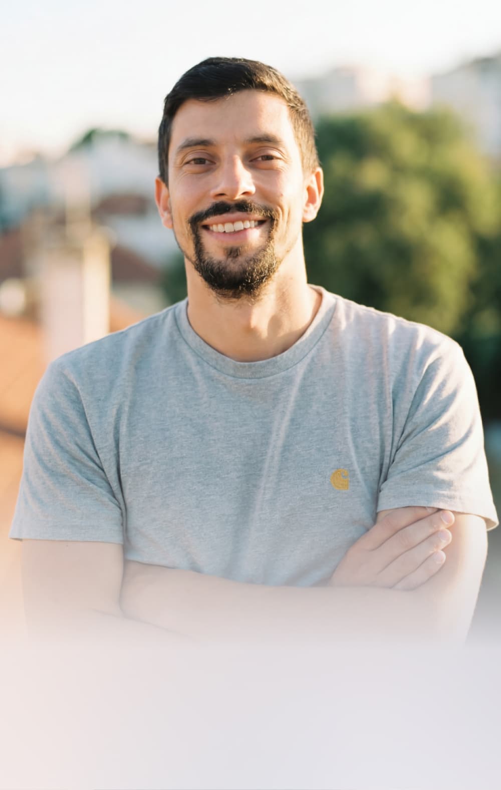 A man with short dark hair and a beard smiles while standing outdoors with his arms crossed, wearing a light gray t-shirt. The background is blurred, showing greenery and rooftops.