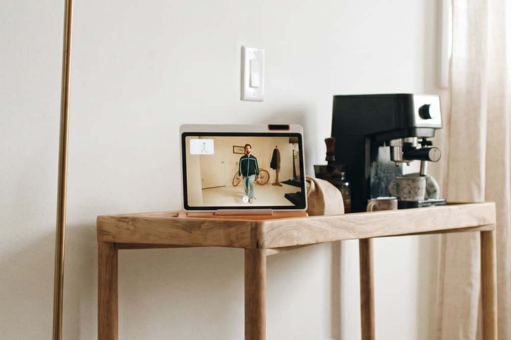 A wooden table holds a tablet displaying a person in workout attire, with a coffee machine and glass items beside it. The setup is against a light-colored wall in a bright room.