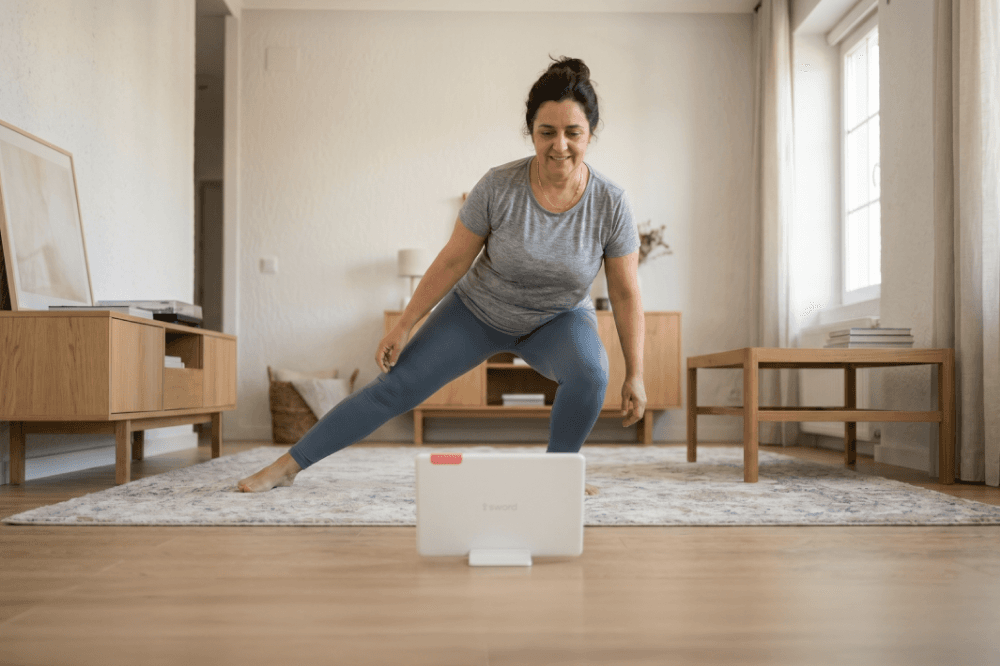 A woman in workout clothes follows an exercise routine on a tablet in her living room, performing a side lunge on a rug in front of modern wooden furniture.