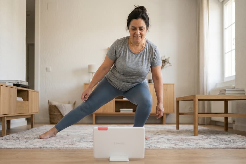 A woman in workout clothes does a side lunge while following an exercise video on a tablet in her living room, which has bright natural light and simple, modern furniture.