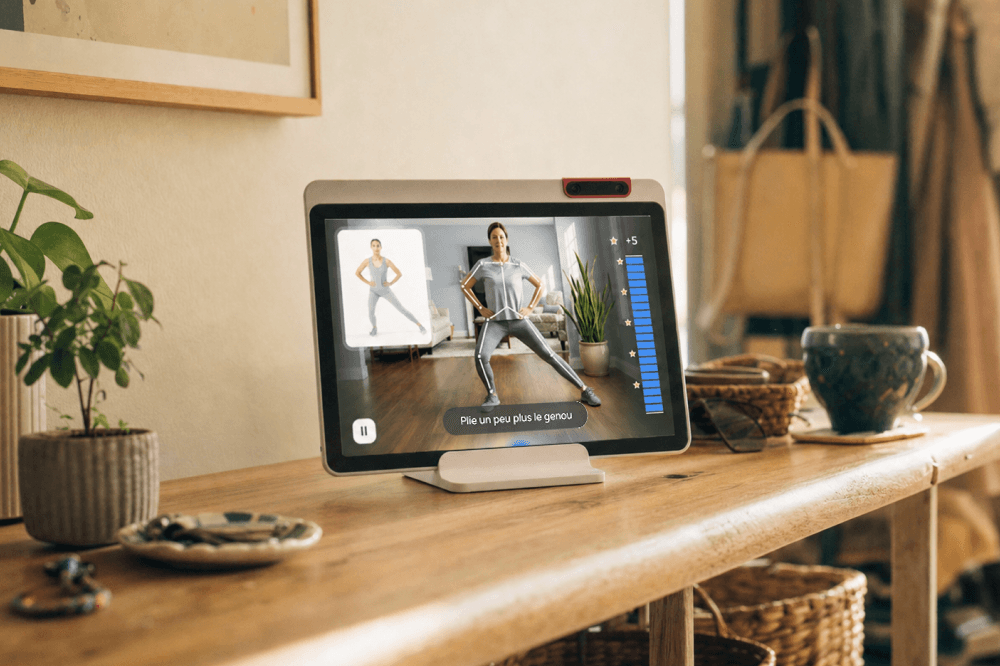 Tablet on a wooden table displays a person doing a workout routine. Plants, baskets, a mug, and other household items are on the table. The screen shows exercise instructions and a progress bar.