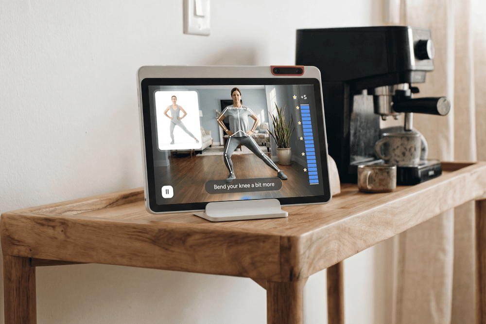 A tablet on a wooden table displays a virtual fitness class with an instructor demonstrating an exercise. The table also holds a coffee machine, coffee grinder, and glass jar.