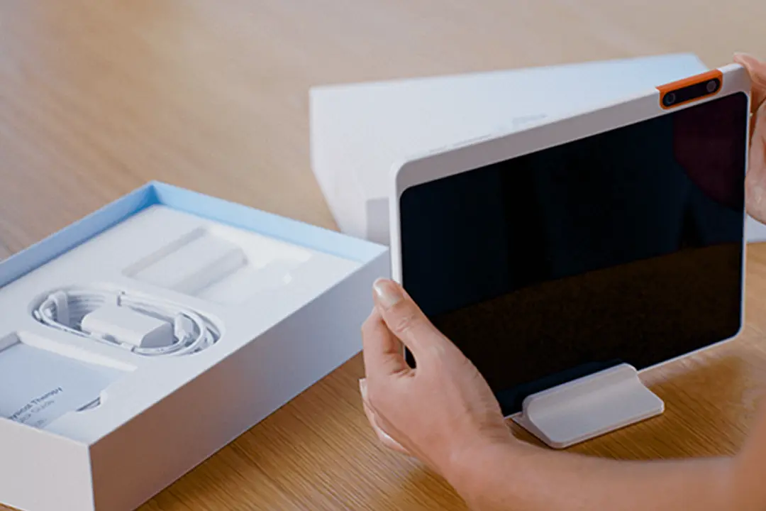 Person holding a tablet with a camera attachment, next to an open box containing a charger and accessories on a wooden table.