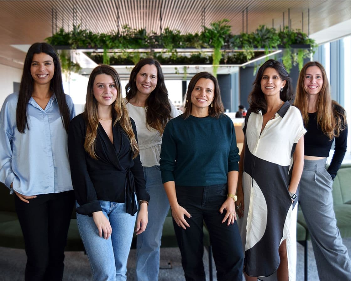 Six women stand together indoors, smiling at the camera. They are dressed in smart-casual attire, with a modern office space and hanging plants in the background.