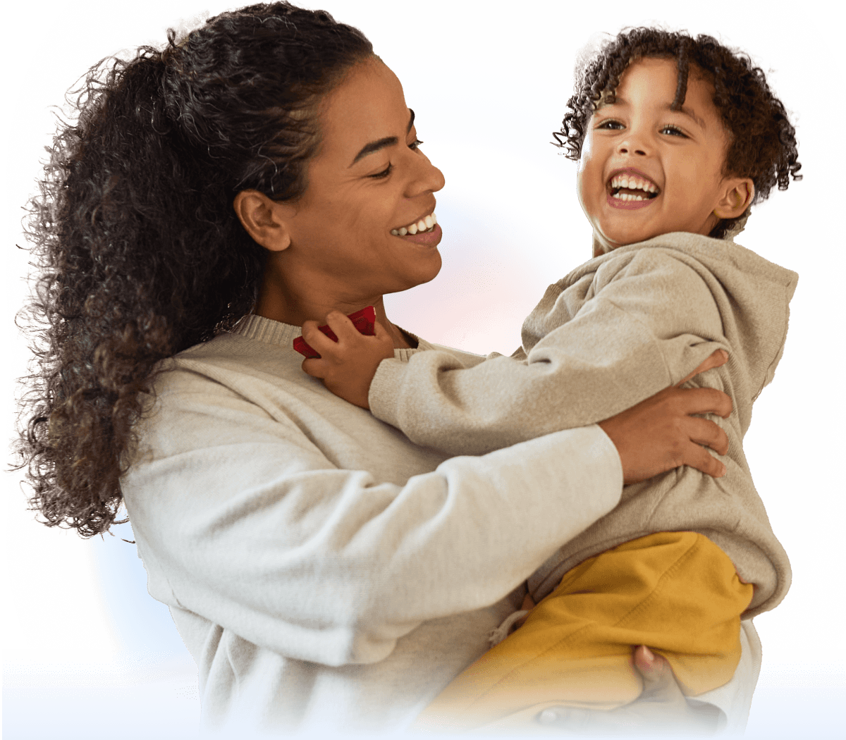A woman with curly hair smiles while holding a laughing young child in her arms. Both are wearing casual, cozy clothes and looking at each other happily against a light background.