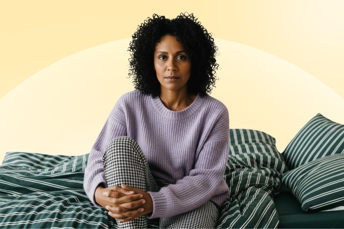 A woman with curly hair sits on a striped bed, wearing a lavender sweater and checkered pants, looking calmly at the camera with a neutral expression against a yellow background.