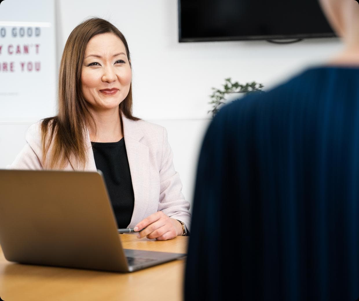 A woman in a light blazer sits at a desk with a laptop, smiling and looking at another person whose back is visible in the foreground. The setting appears to be a professional office.