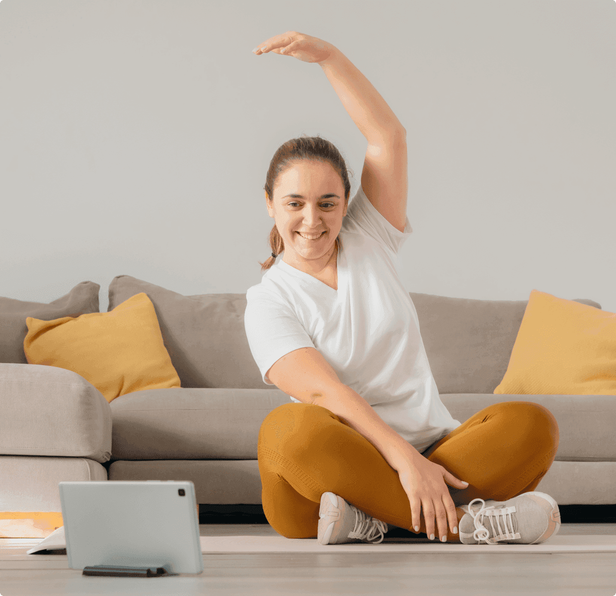 A woman in a white t-shirt and orange leggings sits cross-legged on the floor, stretching her arm overhead and smiling at a tablet in front of her, with a gray couch and yellow pillows in the background.