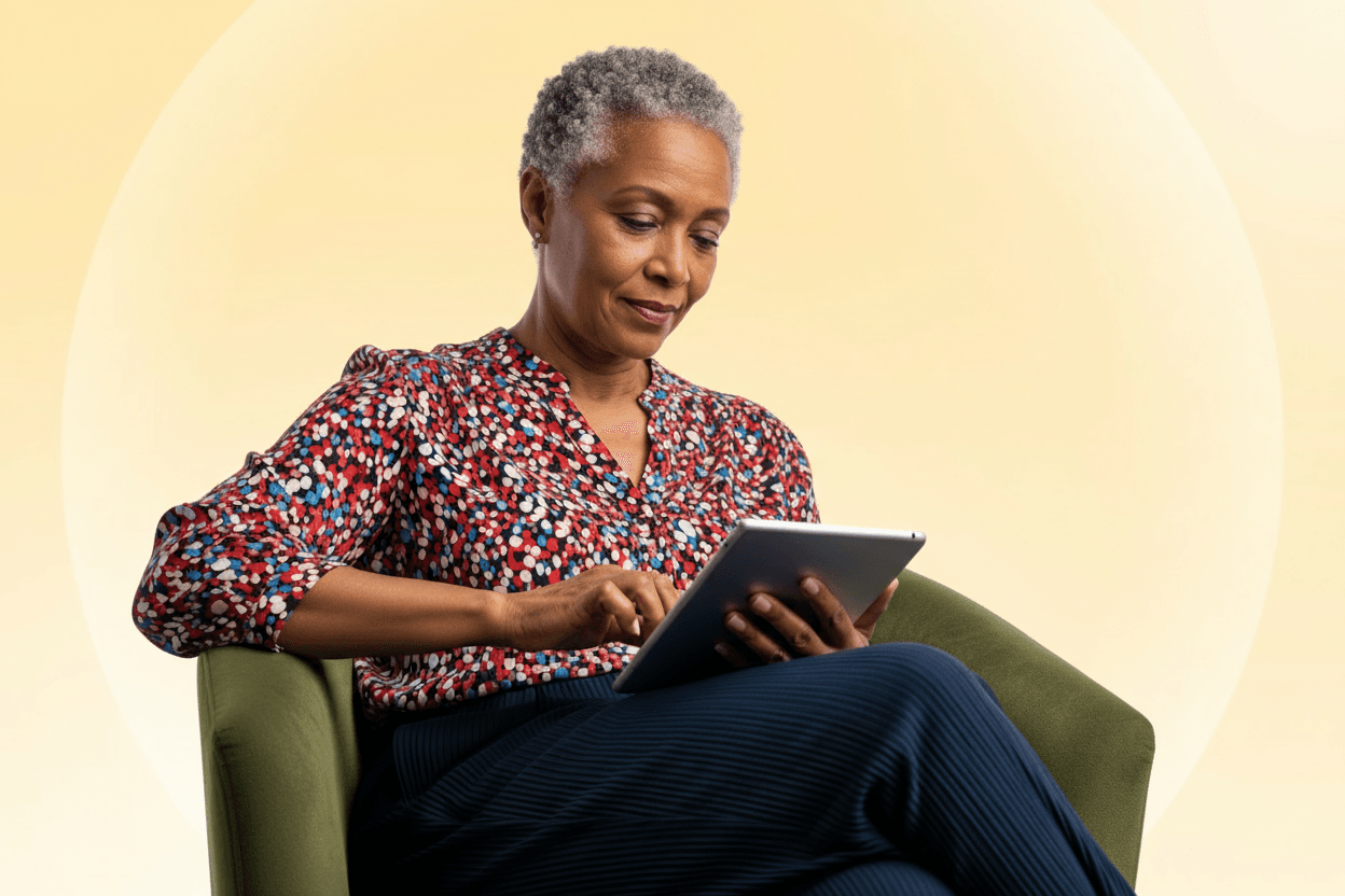 Elderly woman with short gray hair, wearing a floral blouse, sits in a green chair, using a tablet against a soft, light-colored background.
