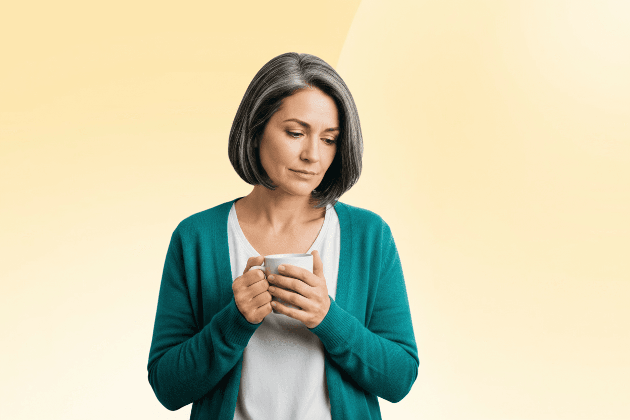 Middle-aged woman with gray bob in teal cardigan holding a white mug, looking down pensively against a soft beige background.