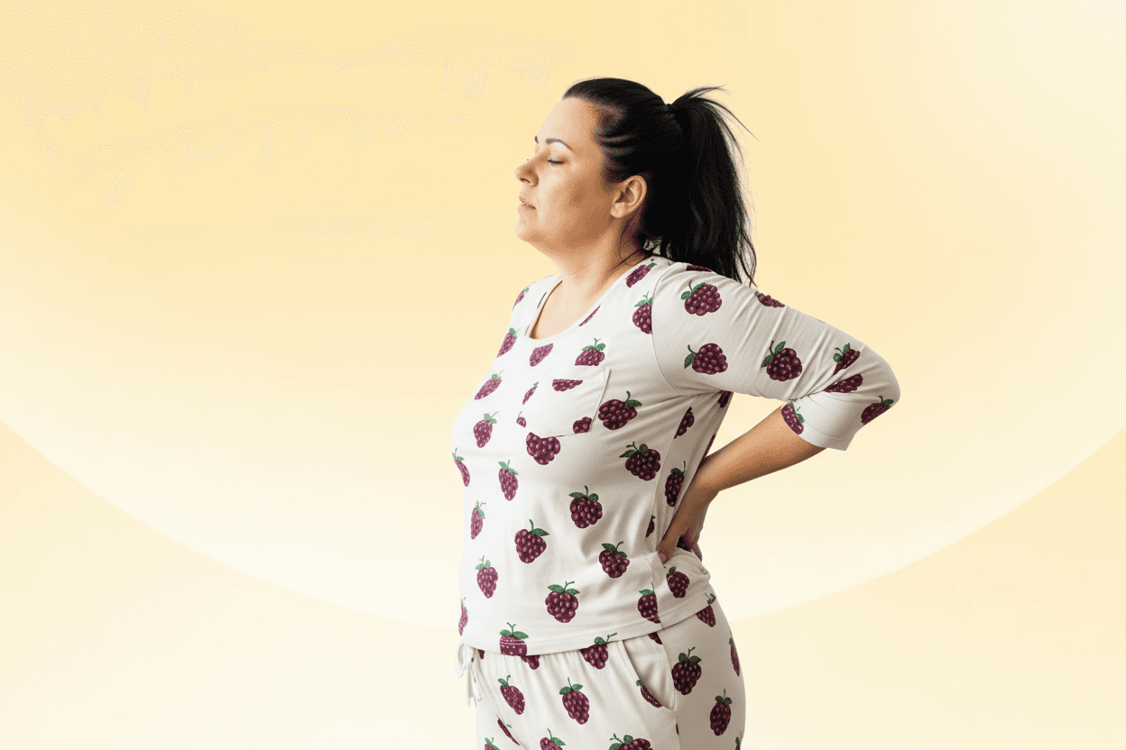Woman in pajamas standing with one hand on her lower back, reacting to back pain after waking up.