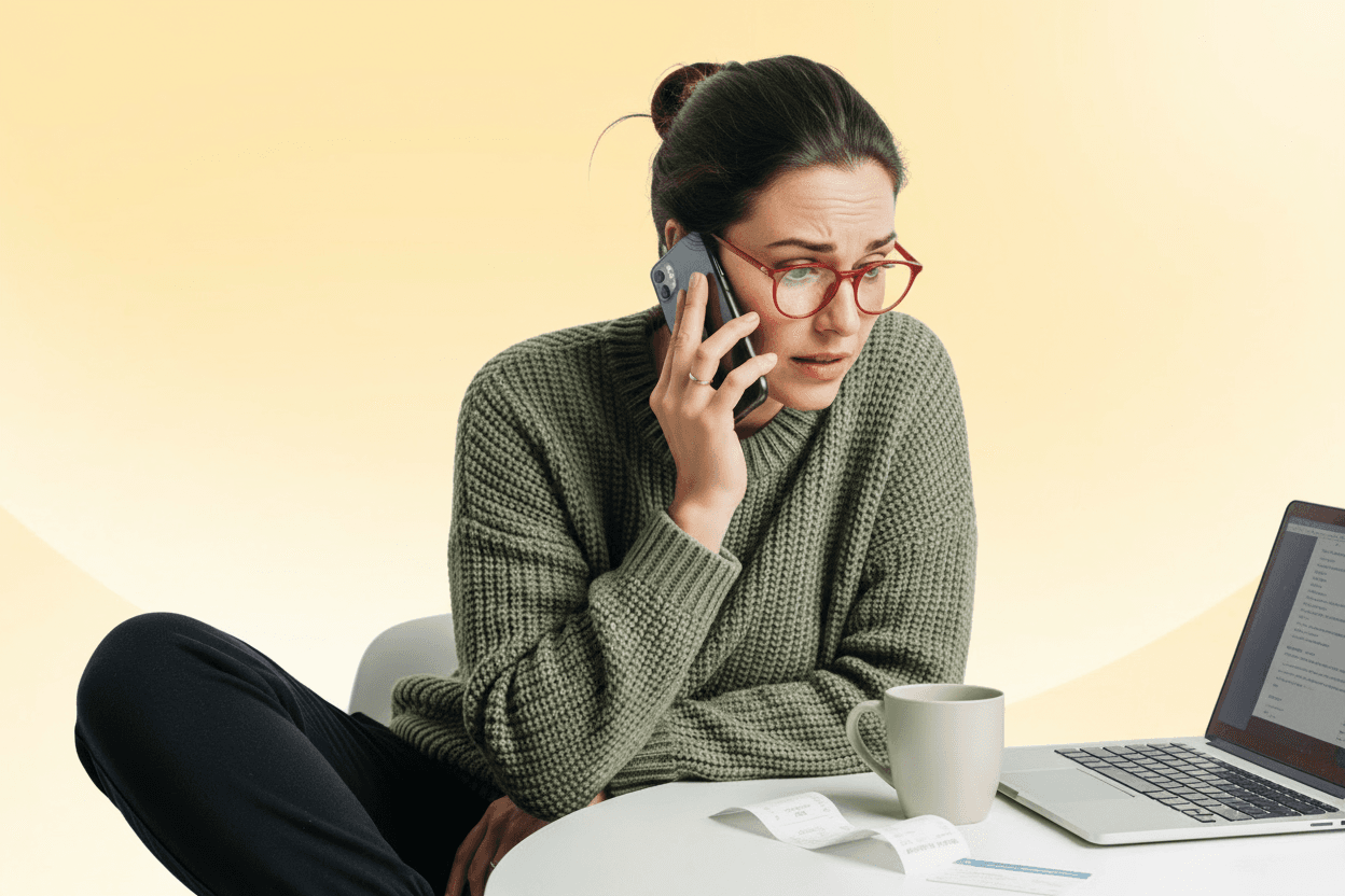 Femme portant un chandail vert et des lunettes rouges, au téléphone, regardant un ordinateur portable avec une tasse de café et des reçus à proximité.