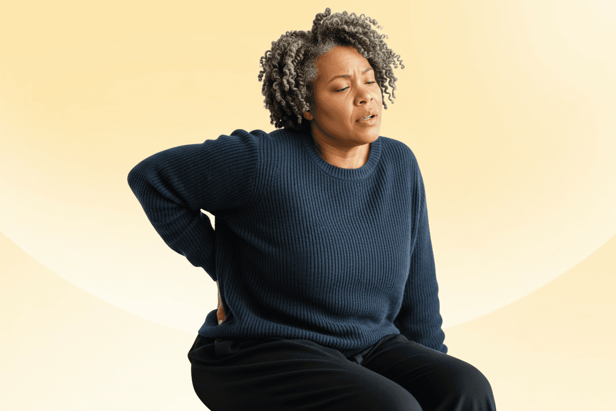 A person with short curly hair and a navy sweater grimaces in pain, holding their lower back against a soft yellow background.