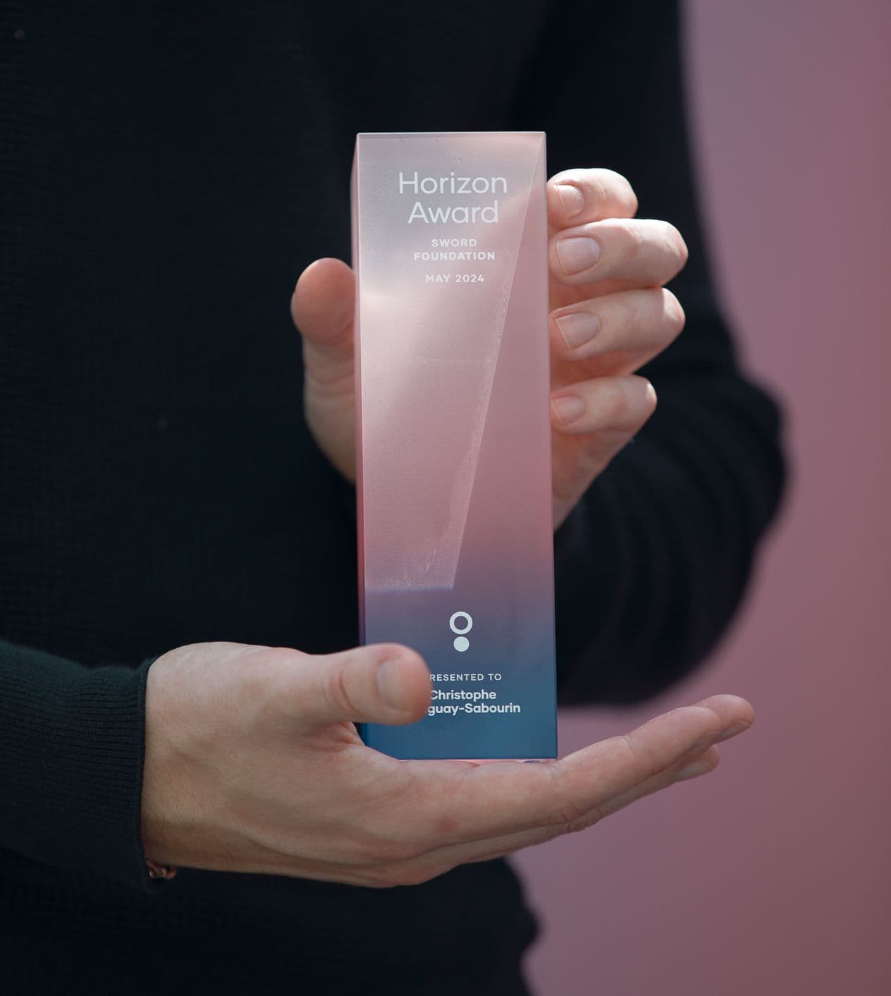 A person in a dark sweater holds a translucent pink and blue rectangular award that reads “Horizon Award, Sword Foundation, May 2024,” with “Presented to Christophe Gauy-Sabourin” engraved at the bottom.