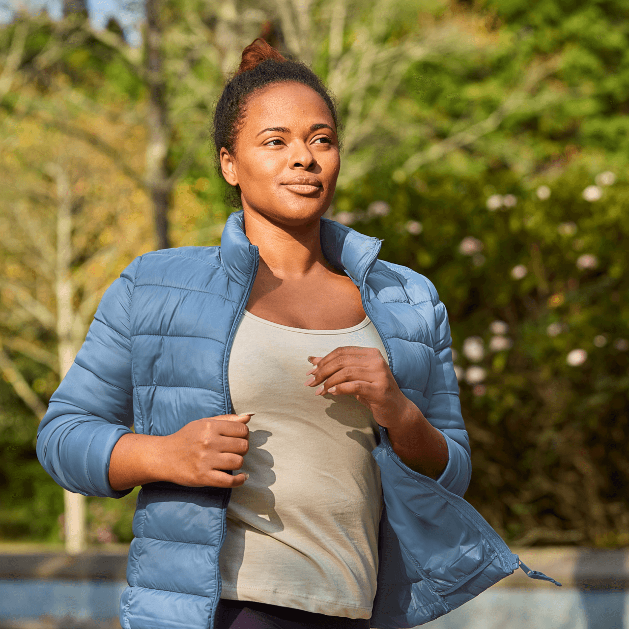 A woman in a blue jacket walks outdoors in a park, surrounded by trees and greenery, under a clear sky.