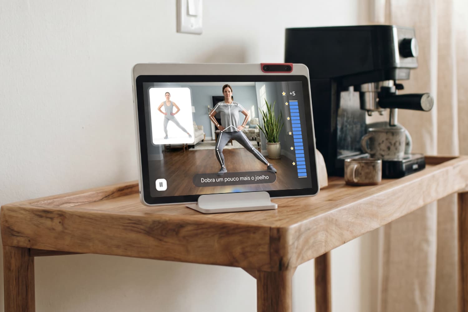 A tablet on a wooden table displays a workout video with a woman exercising. The tablet is next to a coffee machine and grinder in a bright room with curtains.