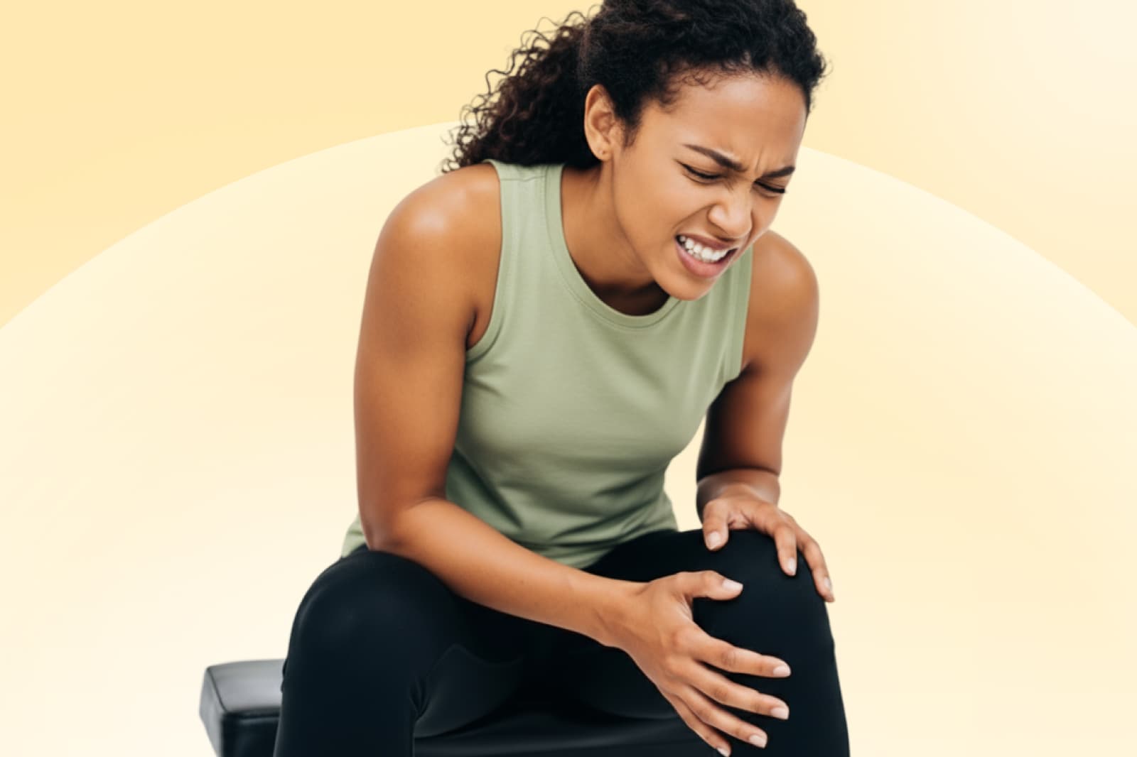 Femme portant un débardeur vert, assise, grimaçant de douleur, tenant son genou, sur un fond jaune doux.