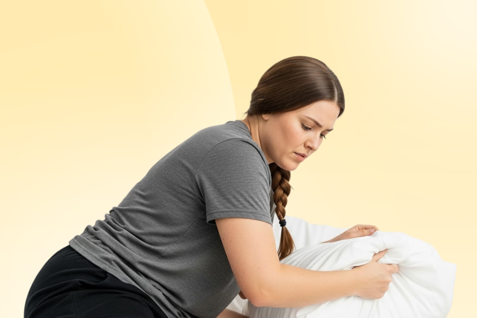 Woman in a grey shirt with a braid adjusts a pillow against a soft yellow background.