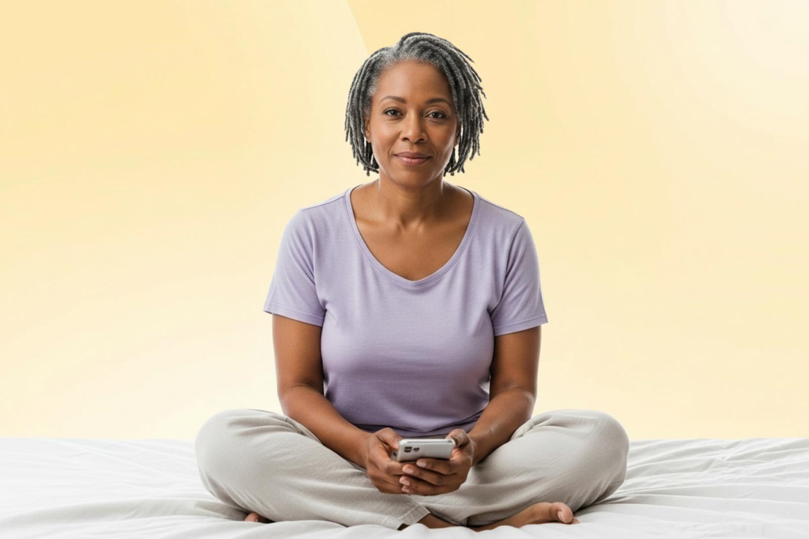 Woman with gray dreadlocks sitting cross-legged on bed, holding smartphone, wearing lavender top and light pants against beige background