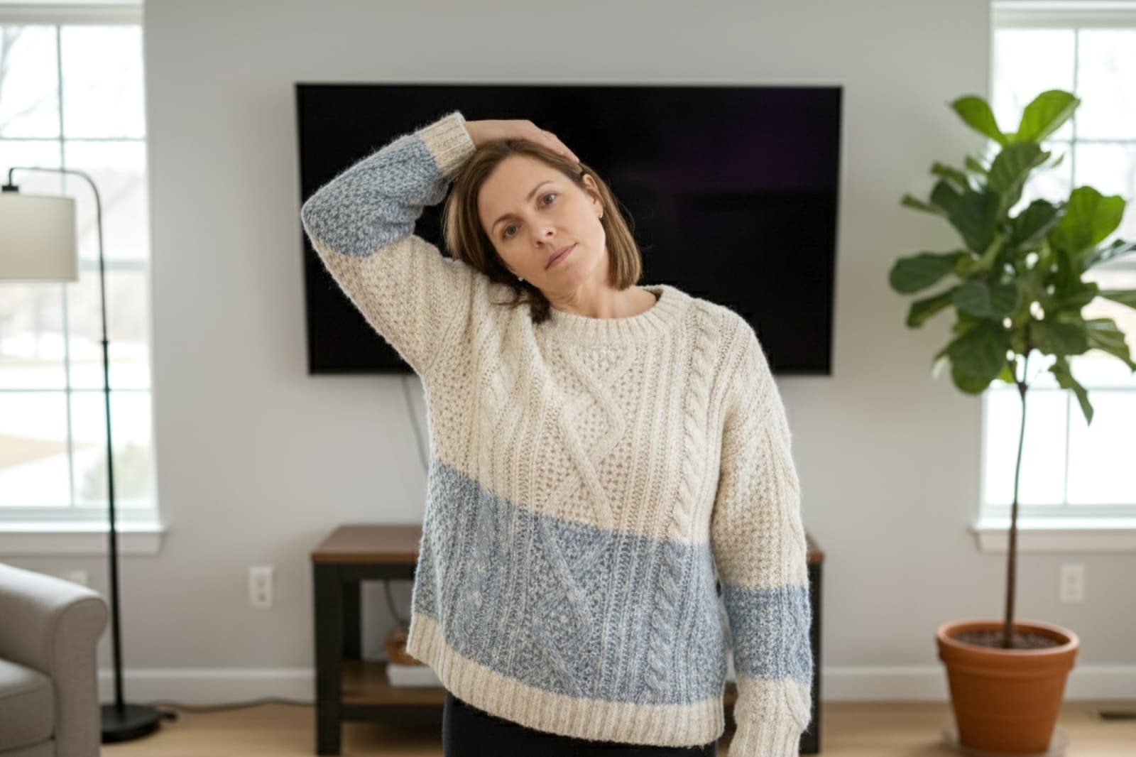 A woman in a cozy sweater tilts her head to the side, stretching her neck in a living room with a TV, windows, and a potted plant.