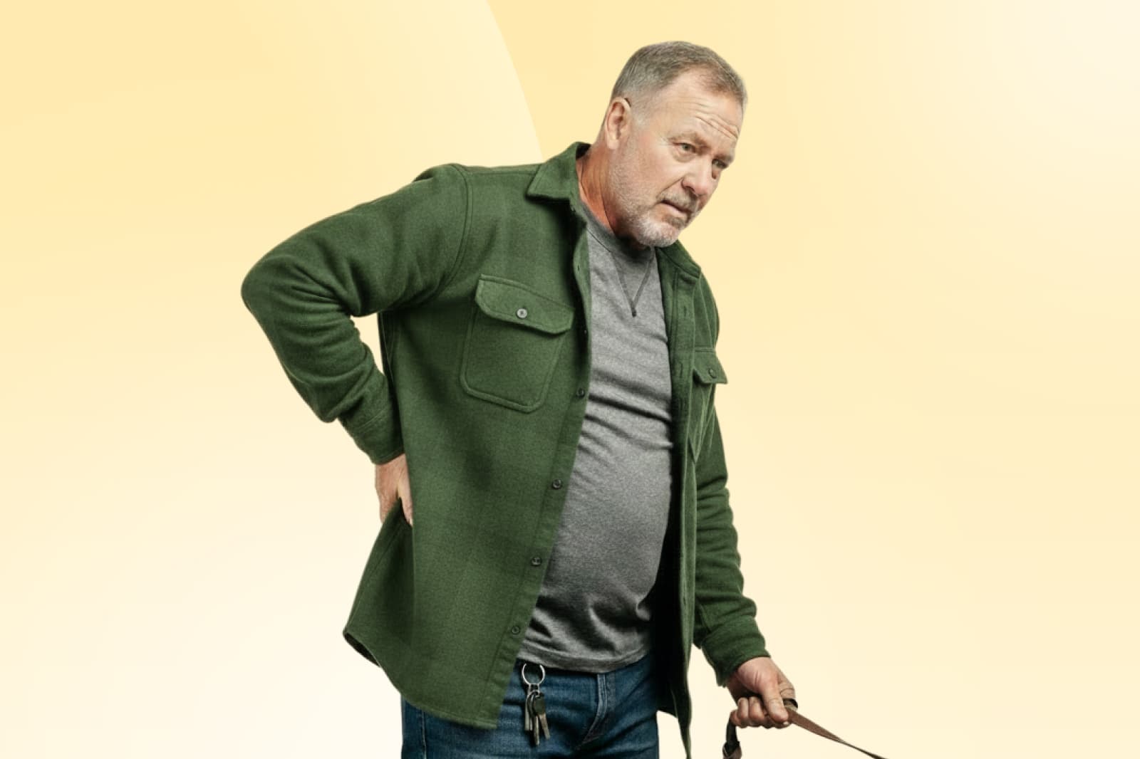 Man in a green jacket holding his back, looking down, against a yellow background. Keys hang from his jeans.