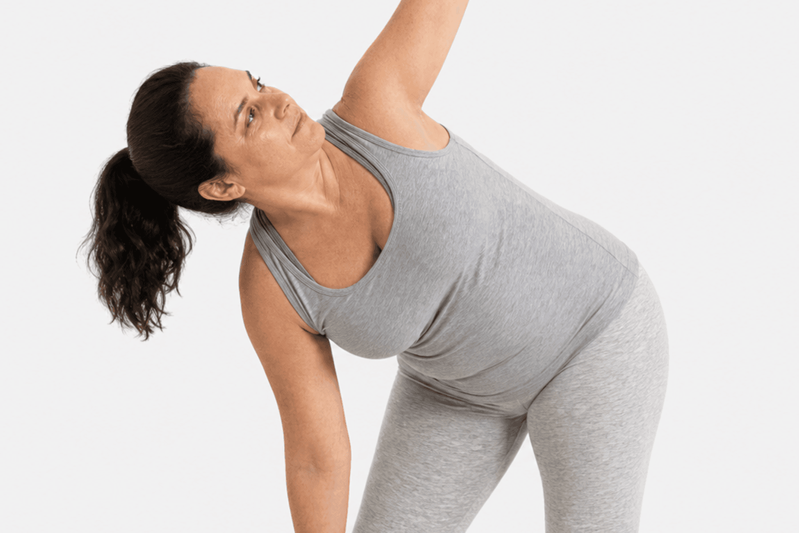 A woman in grey athletic wear performs a side stretch with one arm extended upward and the other reaching down, against a plain white background.