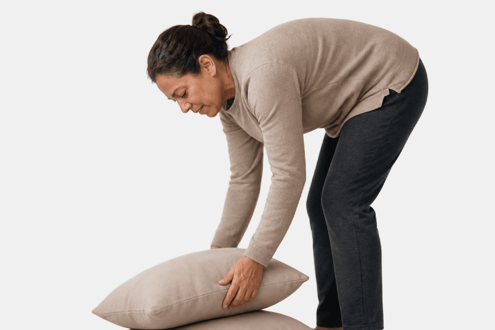 A woman wearing a beige sweater and dark pants bends down to pick up a beige pillow, smiling slightly against a plain white background.