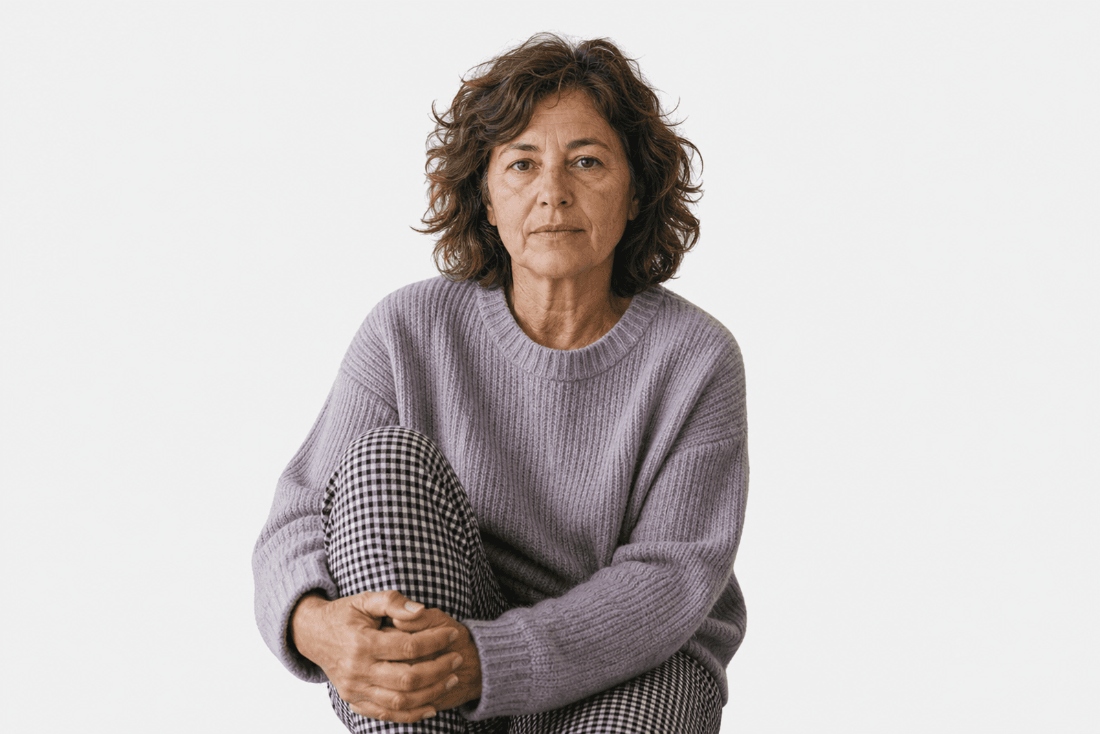 A middle-aged woman with wavy brown hair sits against a plain white background, wearing a light purple sweater and black-and-white checkered pants, looking calmly at the camera.