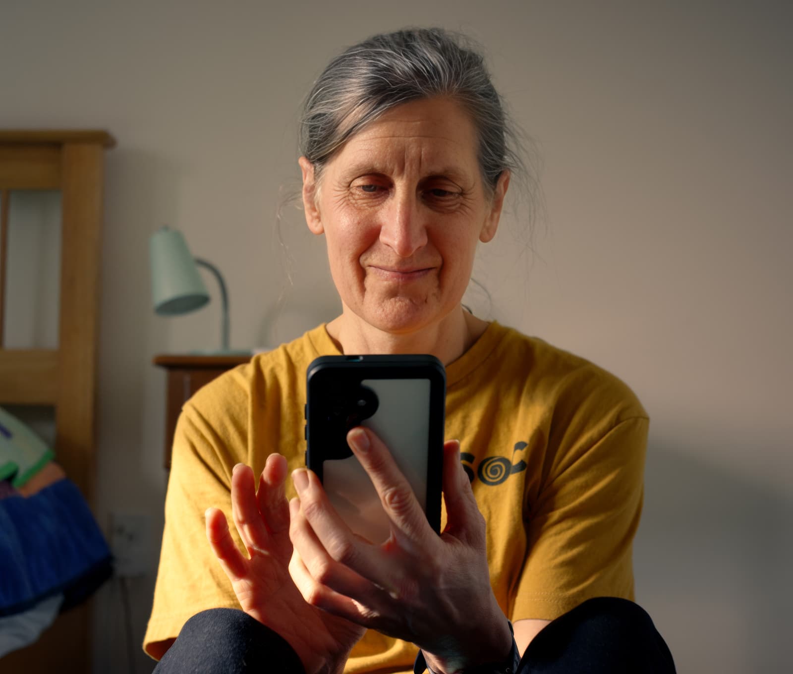 A middle-aged woman with gray hair and a yellow shirt sits indoors, smiling while using a smartphone. Soft natural light illuminates her face, and a lamp and bed are visible in the background.