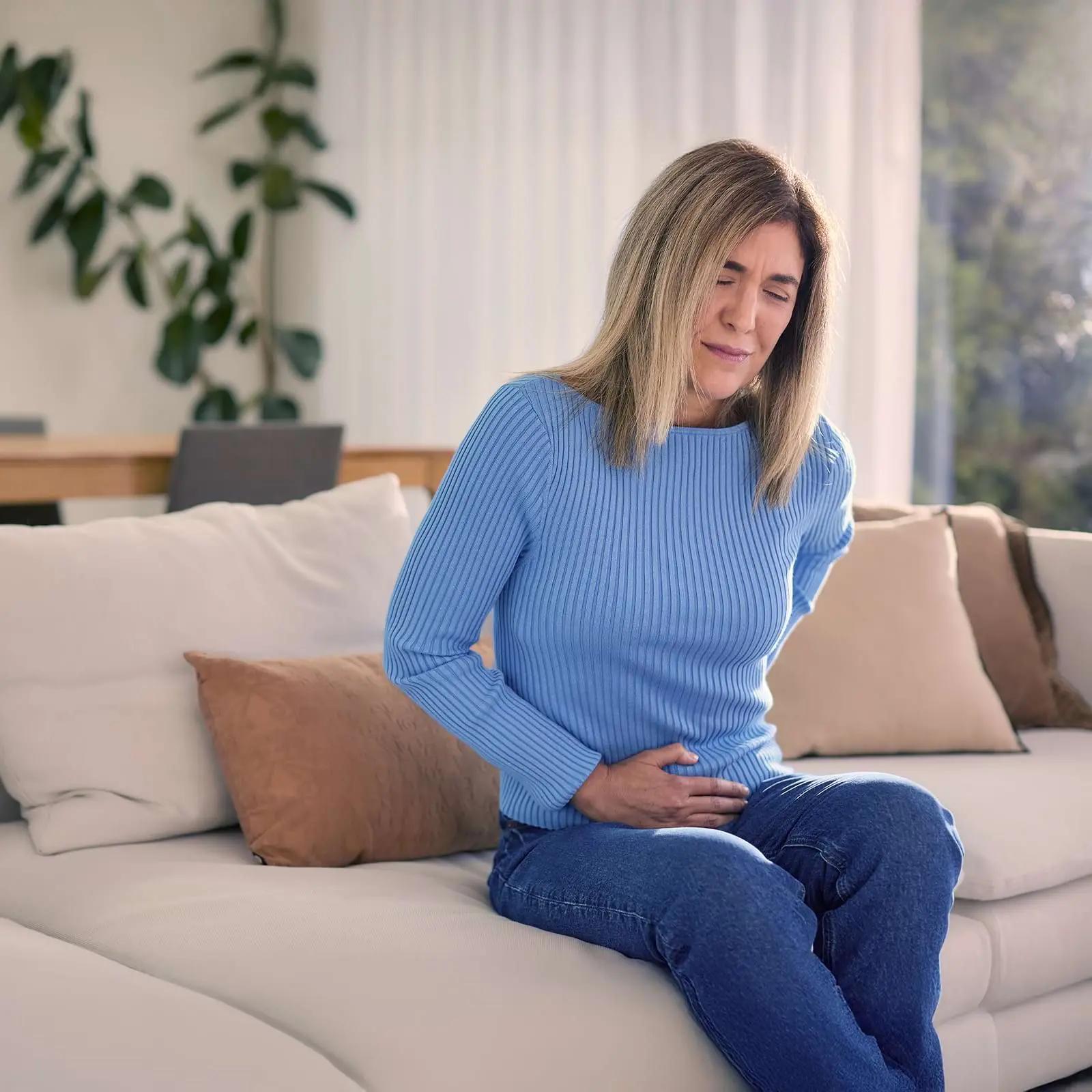 A woman in a blue sweater sits on a sofa, holding her stomach with a pained expression. She appears to be in discomfort.