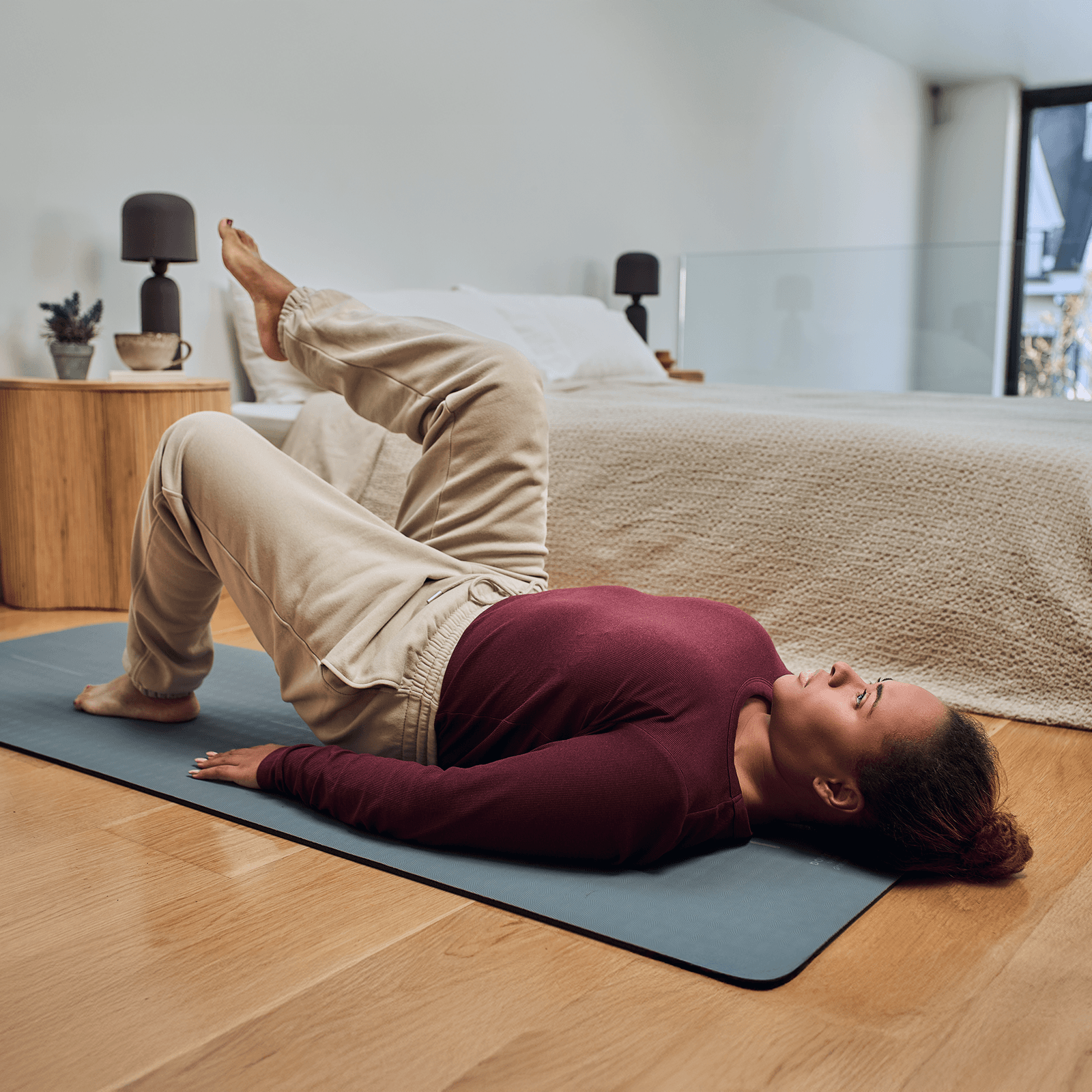 A person in a maroon top and beige pants performs a glute bridge exercise on a mat in a bedroom with wooden furniture.