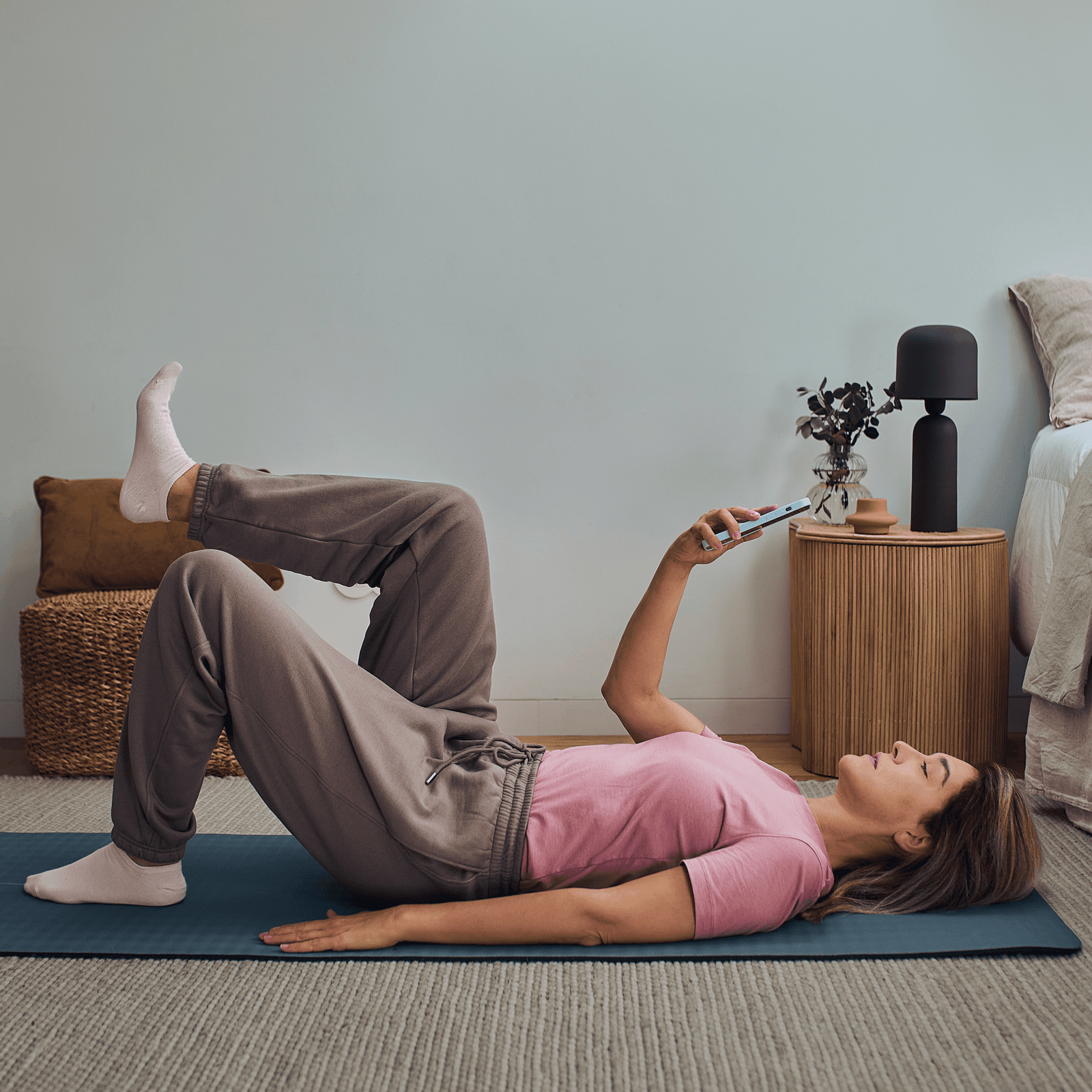 Woman lying on a mat in a relaxed pose, following exercise plan on mobile app. She's wearing a pink shirt and gray pants, with a cozy bedroom setting.